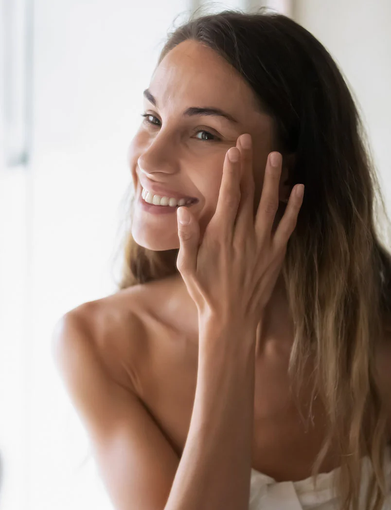 A happy young woman with long brown hair smiles gently while applying a cream or serum to the delicate skin around her eye with her fingers. Her shoulders are bare, suggesting a spa, skincare, or medical aesthetic context, and the image is softly lit, creating a natural and warm atmosphere. - Botox in Greenwood Village, CO
