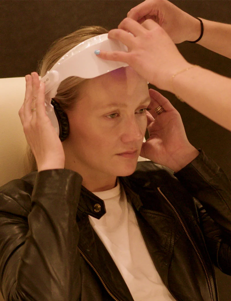 A seated woman with light blonde hair wears a white headband-like device over her forehead. Two hands adjust the device and the surrounding skin. She wears a dark leather jacket over a white t-shirt. The softly lit, blurred background. - Braintap in Greenwood Village, CO
