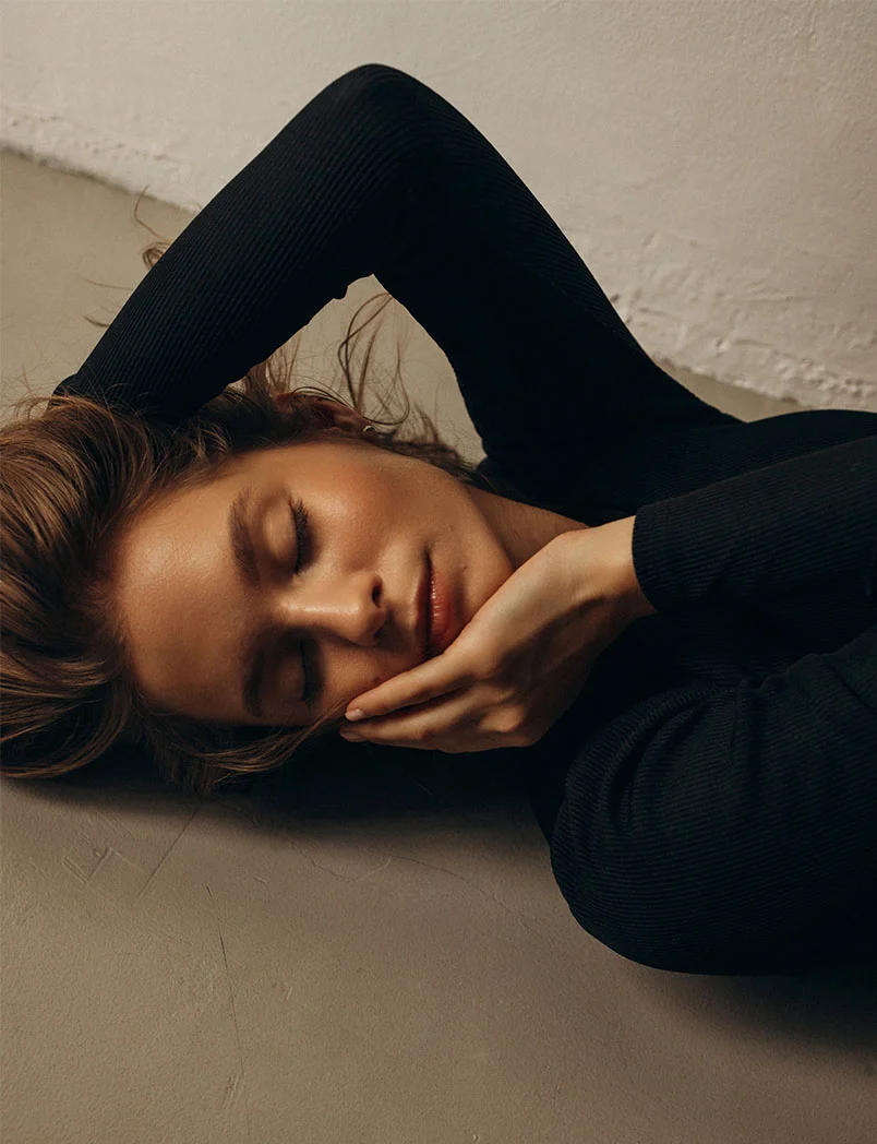 A woman lies on a light taupe floor wearing a black ribbed long-sleeve top. Her brown hair is loose and tousled, one hand rests on her face, the other beside her head. Her eyes are closed with soft, natural makeup. Soft, diffused lighting creates a serene and contemplative mood. - Chemical Peels in Greenwood Village, CO