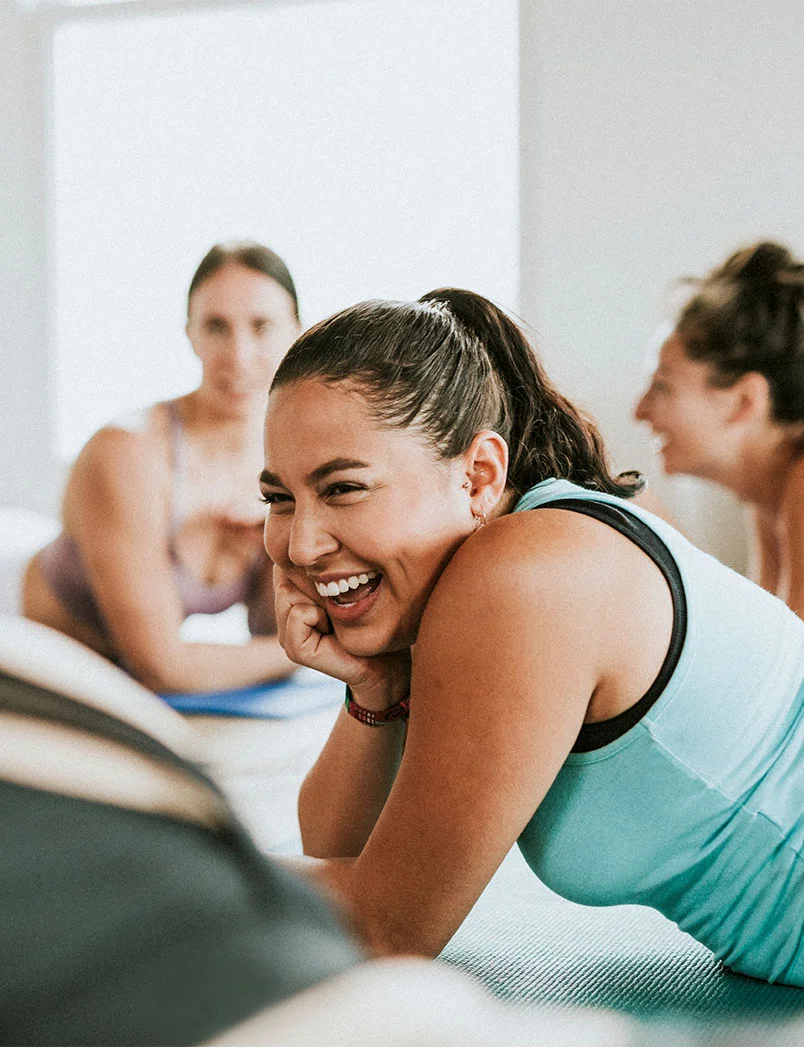 A group of women participate in a fitness class. In the foreground, one woman lies on a mat, laughing with her head tilted back and a hand on her cheek. She has dark brown hair in a ponytail and wears a light teal tank top. Other women are visible in the background, one slightly out of focus, also smiling and engaged. The setting is bright and airy, conveying a joyful, relaxed atmosphere. - Class Offerings in Greenwood Village, CO