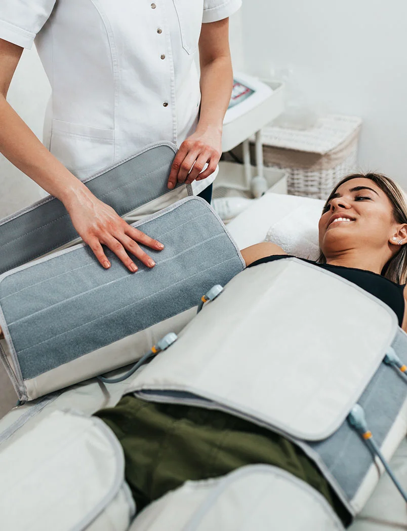 A woman lies on a treatment table wearing olive green clothing, with her torso wrapped in a light gray and white compression therapy device made of multiple panels. A practitioner in a white spa or medical uniform adjusts the device. The woman receiving treatment has a pleasant expression. The setting is a bright spa or clinic room. - Compression Therapy in Greenwood Village, CO