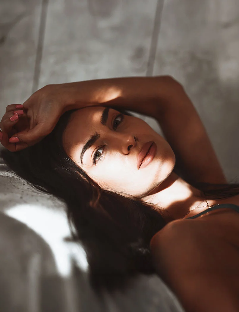A woman lies on a light gray surface, resting her head on one arm with dark hair framing her face. She wears a dark teal lace-trimmed top, her expression contemplative. Soft, diffused lighting highlights her features against a light gray-beige tiled background, creating a calm, intimate atmosphere. - Dermal Fillers in Greenwood Village, CO