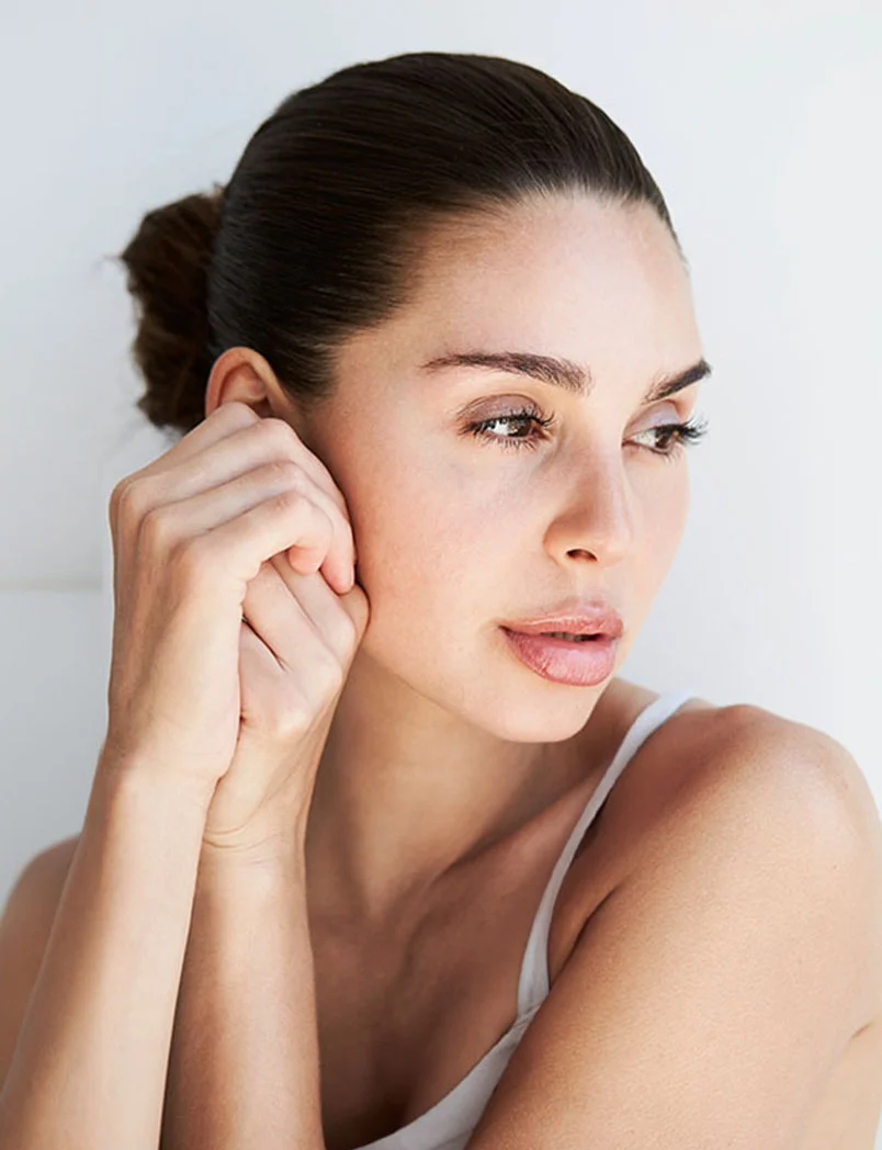 A close-up portrait of a woman with shoulder-length dark brown hair in a low bun. She looks thoughtfully to her right, with smooth, light-toned skin. Her hands gently rest on her cheeks, supporting her head. She wears a plain light-colored tank top against a neutral white background, softly lit to highlight her features. - Diamond Glow Facials in Greenwood Village, CO
