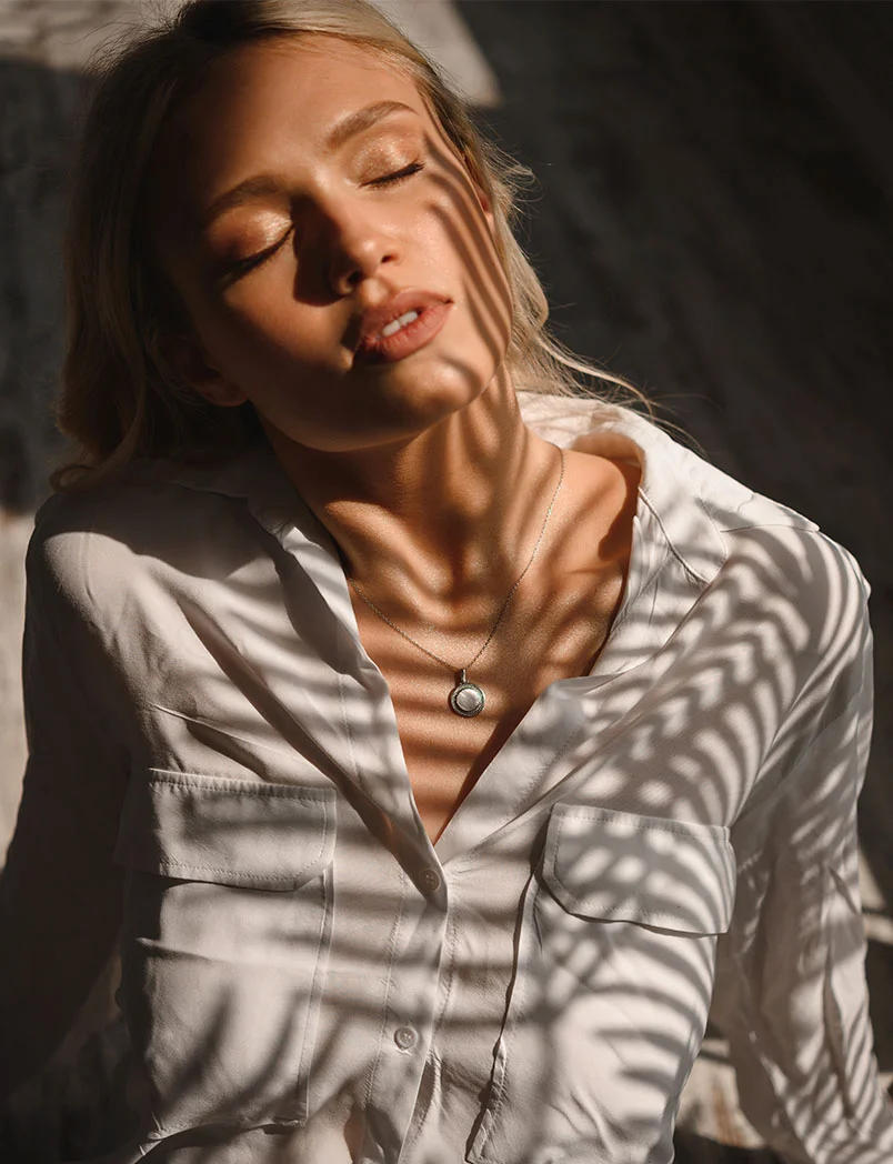 A young blonde woman sits in a dimly lit room with sunlight casting intricate plant-like shadows across her light beige or white button-down shirt, creating delicate, zebra-like patterns. Her eyes are closed, and her expression is serene. A small pendant necklace rests on her chest. The background features a textured light gray and beige surface with darker shadows, highlighting the interplay of light and shadow on her soft features. - Facials in Greenwood Village, CO