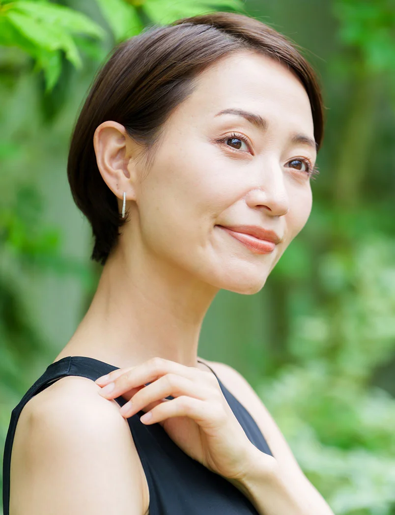 A lovely, middle-aged woman with a short bob haircut and a serene smile, looking off to the side. She is wearing a simple black top and a small, delicate earring. The image is taken outdoors, with a soft, green, leafy background that creates a natural and peaceful atmosphere. - Female BHRT Wellness in Greenwood Village, CO