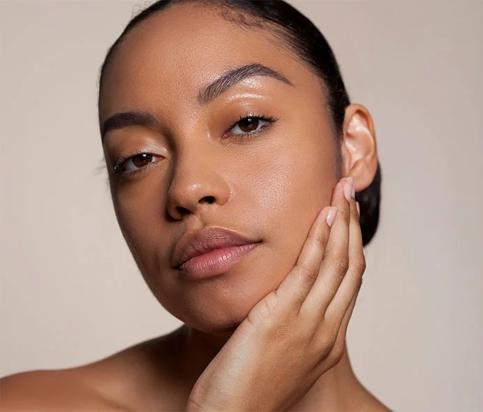 A close-up portrait of a woman with radiant, healthy skin, looking directly at the camera with a gentle and confident expression. Her hair is pulled back, highlighting her smooth skin and natural beauty. She is gently touching her face with one hand, conveying a sense of self-care and grace. - Filler Removal in Greenwood Village, CO