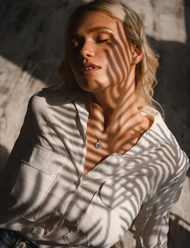 A young blonde woman sits in a dimly lit room with sunlight casting intricate plant-like shadows across her light beige or white button-down shirt, creating delicate, zebra-like patterns. Her eyes are closed, and her expression is serene. A small pendant necklace rests on her chest. The background features a textured light gray and beige surface with darker shadows, highlighting the interplay of light and shadow on her soft features. - Filler Removal in Greenwood Village, CO