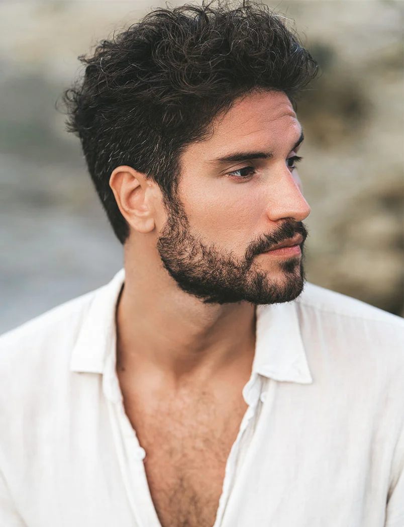 A striking, close-up profile of a handsome man with a full beard and thick, dark, curly hair. He is looking off to the side with a serious, thoughtful expression, wearing a light-colored shirt unbuttoned at the top. The background is slightly blurred, giving the image a soft, focused feel. The man's hair and beard are prominent, suggesting a context related to hair care or hair restoration. - Hair Restoration with Exosomes in Greenwood Village, CO