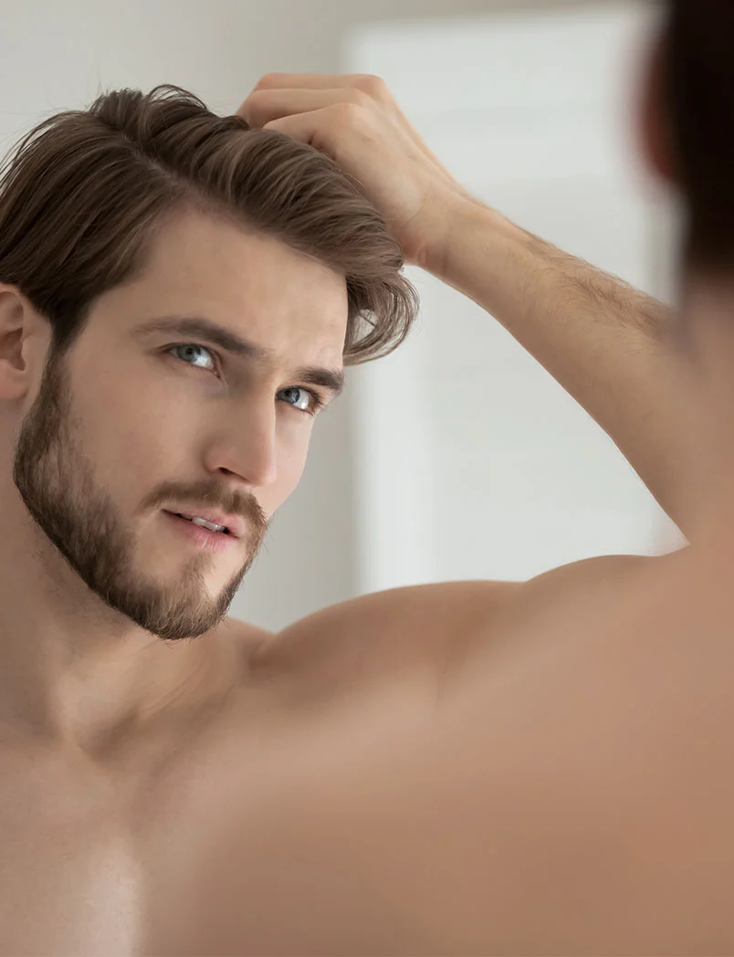 A man with a neatly trimmed beard and brown hair is looking directly at the camera, with one hand running through his hair. His blue eyes are prominent, and his bare shoulder and chest are visible, suggesting a close-up, personal shot. The background is a soft, light color. - Hair Restoration with PRF in Greenwood Village, CO