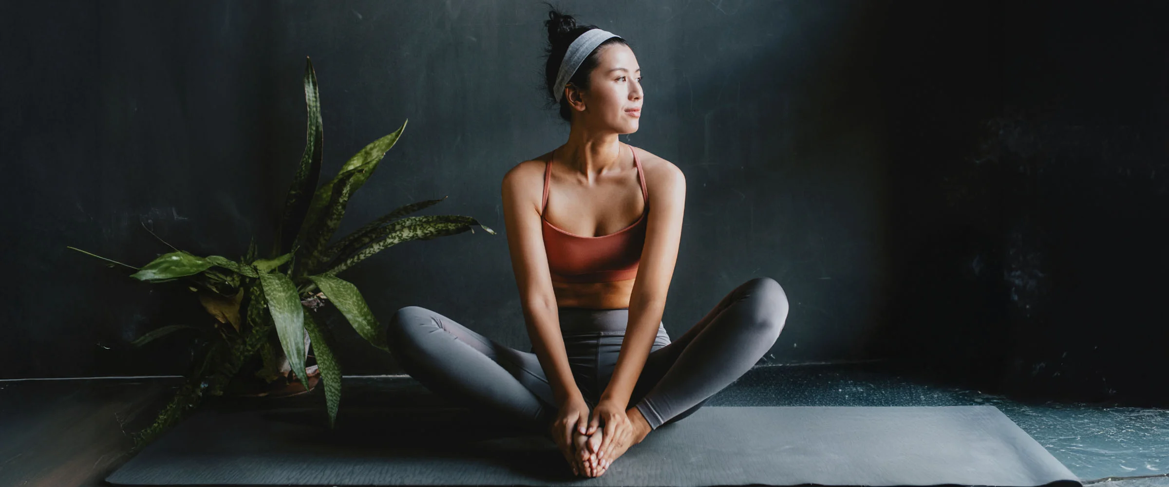 A serene young woman with her hair in a bun and a headband, wearing an orange sports bra and gray leggings, sits in a butterfly stretch on a yoga mat. She is looking off to the side with a calm expression. A large potted plant sits to her left, and the background is a dark, textured wall, creating a simple, peaceful, and focused atmosphere. - Inbody in Greenwood Village, CO