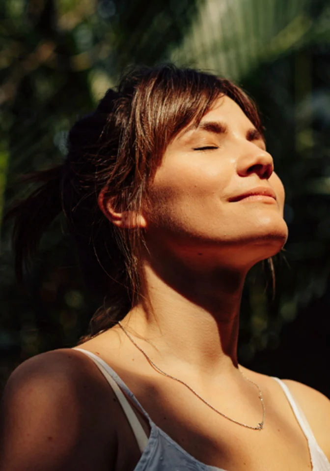 A serene woman with her eyes closed, basking in the sun. She has light brown hair tied back and is wearing a white tank top with a delicate silver necklace. The background is a soft focus of green foliage. - Laser Skin Rejuvenation in Greenwood Village, CO