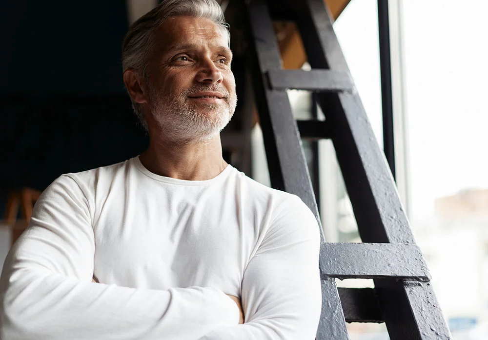 A handsome, middle-aged man with graying hair and a beard, wearing a white long-sleeved shirt, stands with his arms crossed and looks off into the distance with a gentle smile. He is positioned indoors next to a dark, metal structure and a large window, with natural light illuminating his face from the right. The image has a soft, inviting tone, suggesting wellness, contentment, and thoughtfulness. - Male BHRT Wellness in Greenwood Village, CO