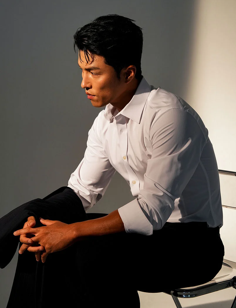 A well-dressed man with dark hair is sitting on a chair, looking off to the side with a pensive expression. He is wearing a crisp white button-down shirt and black trousers, with a dark jacket draped over his arm. The lighting creates a strong contrast between the light and shadow on his face and the white wall behind him. - Male BHRT Wellness in Greenwood Village, CO