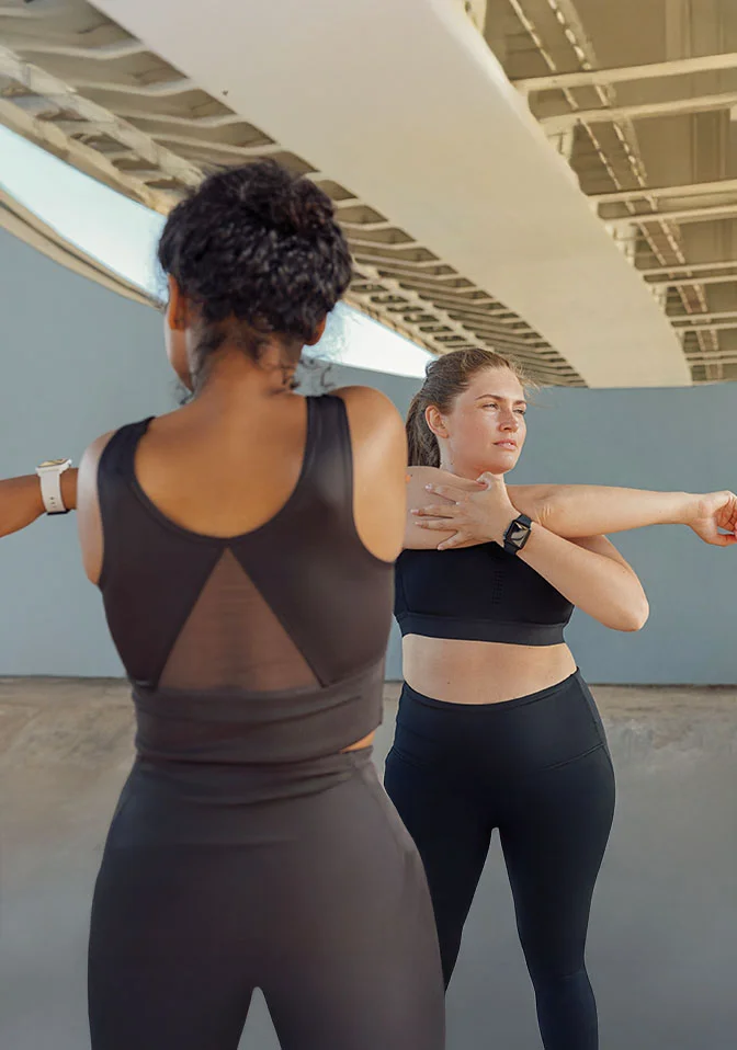 Two women in black workout clothes are stretching their arms. The woman on the left has her back to the camera and a bun. The woman on the right is facing sideways and holding her arm in a stretch. They are standing outdoors under a bridge or overpass. - Medical Weightloss in Greenwood Village, CO