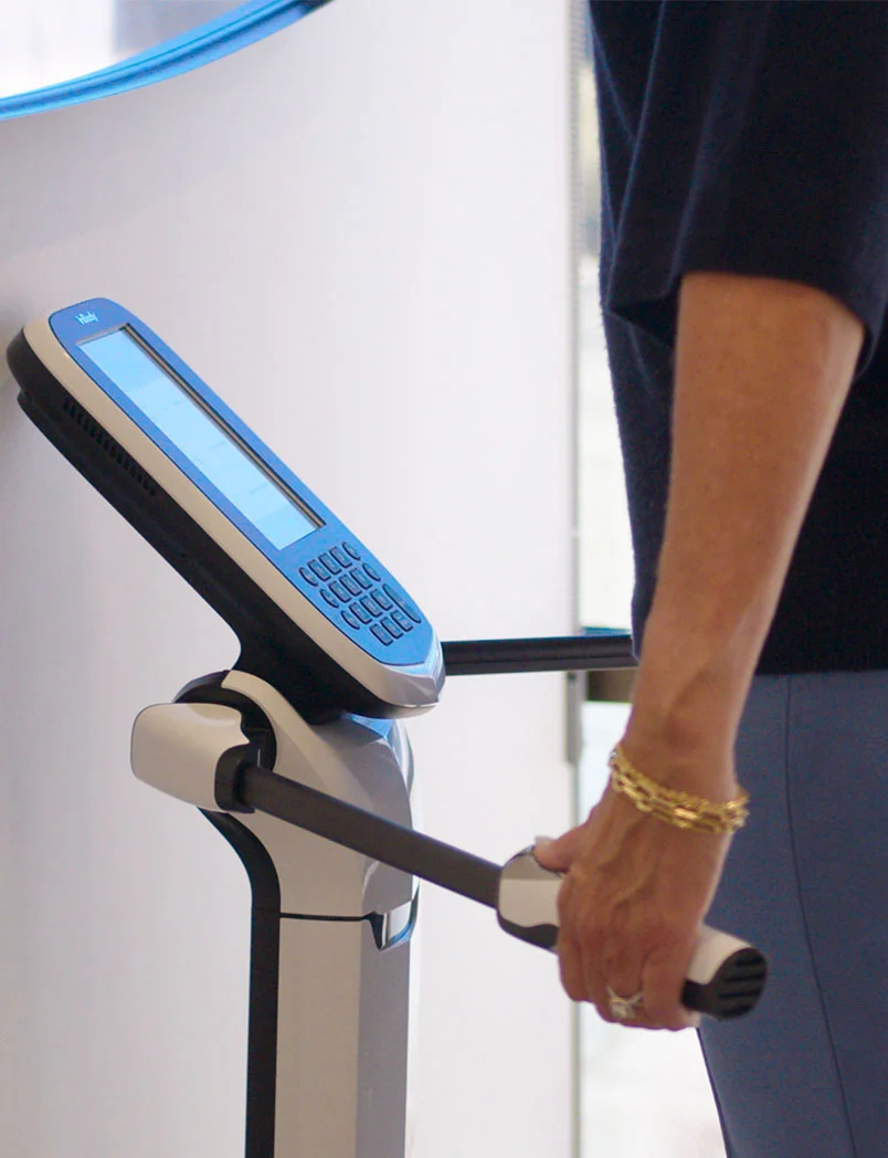 A person's arm, adorned with gold bracelets, is holding the handle of a body composition analysis machine. The machine has a blue screen and keypad, and the person is wearing a dark shirt. - Medical Weightloss in Greenwood Village, CO