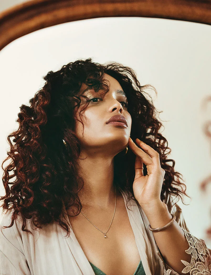 A woman with dark, curly hair is looking at her reflection in a mirror. She is gently touching her chin with one hand. She is wearing a light-colored robe with lace trim, a silver necklace, and a bracelet. - Microneedling in Greenwood Village, CO