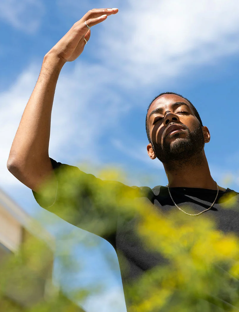 Low-angle close-up of a dark-skinned man with his arm raised, hand shading his eyes. His eyes are closed, and a soft shadow falls across his face from sunlight above. The background shows a bright, partly cloudy blue sky and blurred yellow-green vegetation below. A simple chain necklace rests on his chest over a dark shirt. - Peptide Injections in Greenwood Village, CO
