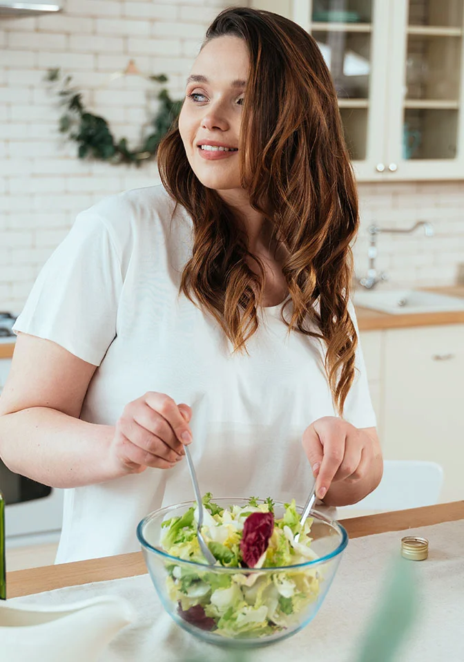 A woman with shoulder-length wavy brown hair smiles gently, looking to her right. She wears a loose white t-shirt and is tossing a salad of green and red leafy greens in a clear glass bowl. The kitchen background features a light beige brick wall, white cabinets, a light countertop with a tablecloth, and leafy green sprigs on a wall decoration. - Personal Training and Nutrition in Greenwood Village, CO