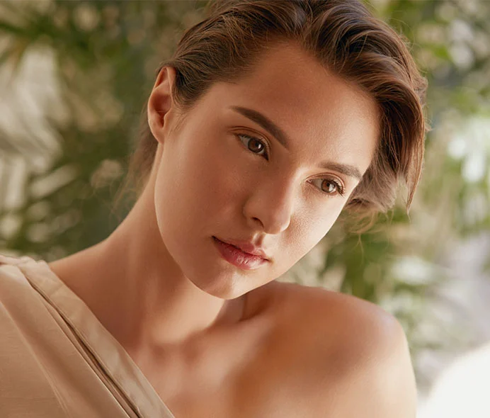Close-up of a young woman with a tousled short bob of light brown hair and smooth, light tan skin. She looks downward and slightly left with a thoughtful expression and soft gaze. She wears a light beige off-the-shoulder top. The blurred green background suggests a garden, with soft natural light highlighting her face and upper body. - PRF Injections in Greenwood Village, CO