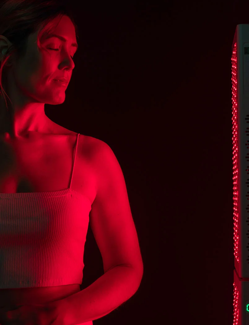A woman with light brown hair is shown in profile with eyes closed and a serene expression. She wears a light ribbed tank top, bathed in vibrant deep red light. To her right, a tall rectangular device with rows of red LEDs and a small green light contrasts against a black background, highlighting the woman and the light therapy device. - Red Light Therapy in Greenwood Village, CO