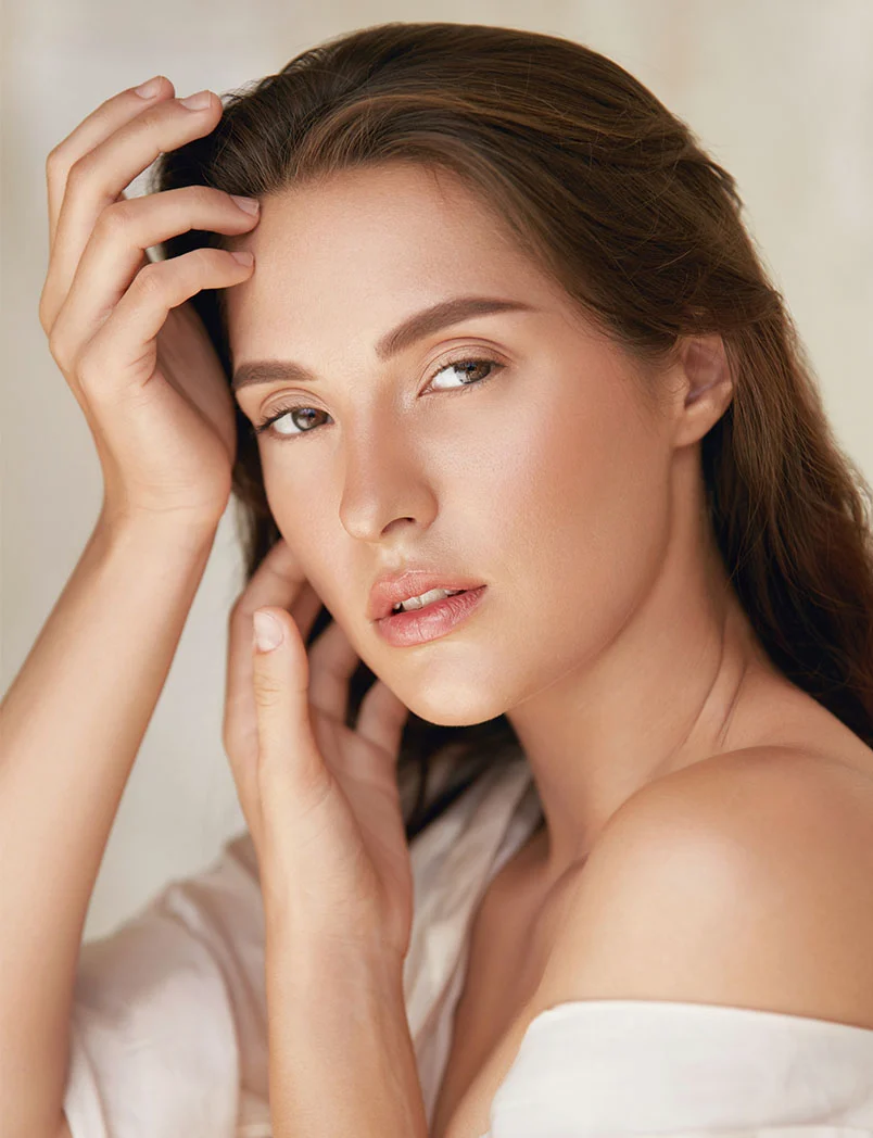 Close-up portrait of a woman with long dark brown hair, light beige-toned skin, and large brown eyes. She wears light cream off-the-shoulder clothing, with one hand above her forehead and the other near her jawline. Soft, diffused lighting and a light beige background highlight her serene, elegant expression. - Sculptra in Greenwood Village, CO