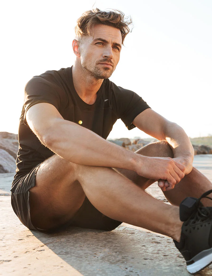 A light-skinned man with tousled light brown hair sits on a light tan stone surface, wearing dark gray athletic shorts and a matching t-shirt. His legs are bent, hands resting on them, and he gazes to his right with a contemplative expression. Black athletic shoes are visible. Warm, low sunlight suggests an outdoor coastal setting. - Wellness Injections in Greenwood Village, CO